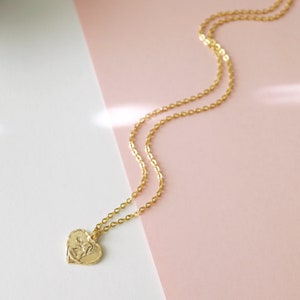 May include: A gold chain necklace with a heart-shaped pendant featuring a detailed design. The necklace is on a pink and white background.