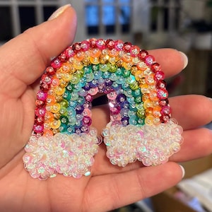 May include: A rainbow-colored sequin patch with white sequin clouds. The rainbow is made of small, round sequins in shades of red, orange, yellow, green, blue, indigo, and violet. The clouds are made of white, iridescent sequins.