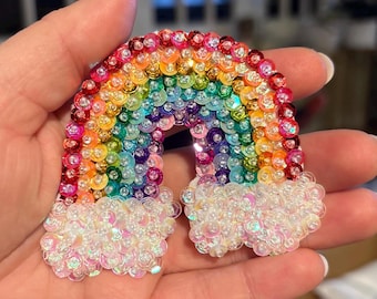 Puede incluir: Un parche de lentejuelas arcoíris con nubes de lentejuelas blancas. El arcoíris está hecho de pequeñas lentejuelas redondas en tonos de rojo, naranja, amarillo, verde, azul, añil y violeta. Las nubes están hechas de lentejuelas blancas iridiscentes.