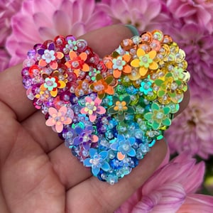 May include: A heart-shaped decorative item, crafted with colorful sequins and small flower embellishments. The design features a rainbow of colors, including red, orange, yellow, green, blue, and purple. The item is held in a hand, with a blurred floral background.