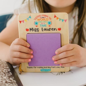 May include: A wooden teacher appreciation gift with a purple sticky note pad. The text on the gift reads "Teachers change the world one child at a time" and "Miss Lauren". The gift is held by a child at a desk with school supplies.