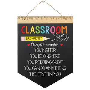 May include: Black chalkboard classroom rules sign with a rainbow colored word "CLASSROOM" and white text that reads "MRS. MARTINEZ Rules Always Remember YOU MATTER YOU BELONG HERE YOU'RE DOING GREAT YOU CAN DO ANYTHING I BELIEVE IN YOU".