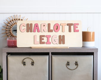 May include: Wooden name puzzle with the name "CHARLOTTE LEIGH" in pink and brown letters. The puzzle sits on a wooden stand. The letters are in a variety of pink and brown shades. The puzzle is on a gray shelf with two storage bins.