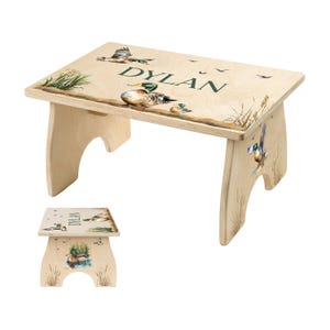 May include: A wooden step stool with a duck theme. The stool is light brown with a painted design of ducks, reeds, and the name "DYLAN" in green. The stool has a rectangular top and curved legs.