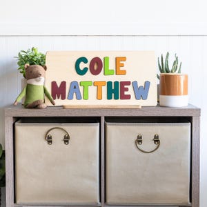May include: A wooden name puzzle with the name "COLE MATTHEW" in colorful, raised letters. The puzzle sits on a gray shelf with two beige storage bins below. A stuffed animal and potted plants are on the shelf.
