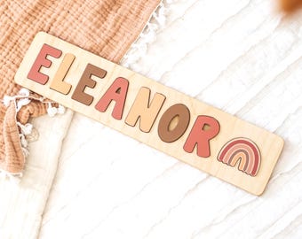 May include: Wooden name puzzle spelling "ELEANOR" in various shades of pink, brown, and beige. A small rainbow graphic is at the end. The puzzle is on a light-colored wooden base.
