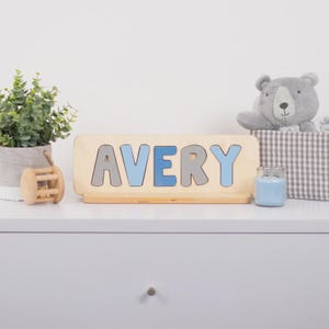 A wooden nameplate with the name "AVERY" on it, placed on a white dresser next to a potted plant and a stuffed teddy bear.