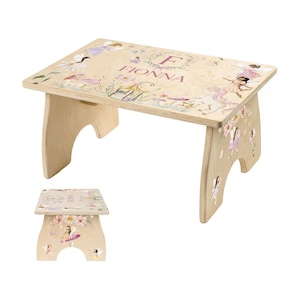 May include: A small, wooden step stool with a light wood finish. The top is decorated with a floral and fairy design, with the name "FIONNA" printed in pink. The stool has a rectangular top and curved legs.