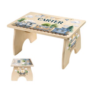 May include: A light-colored wooden step stool featuring a watercolor train illustration and the name CARTER printed on the top surface. The side panel displays a smaller train graphic with clouds and trees, showcasing a consistent design theme.
