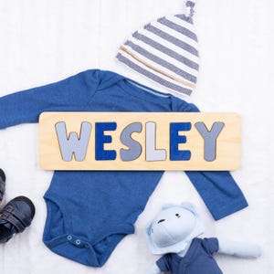 May include: A wooden puzzle spelling "WESLEY" in blue and gray letters, resting on a blue onesie. A striped hat, a plush toy, and baby shoes are also in the image. The items are arranged on a white surface.