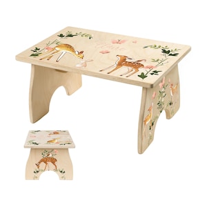 May include: A wooden step stool with a light wood finish. The top surface is decorated with illustrations of deer, a rabbit, flowers, and the name "Claire". The legs also feature deer and floral designs. The stool is designed for children.