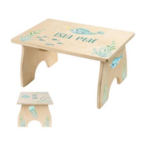 May include: A light wood children's step stool with a marine theme. The stool features a narwhal, fish, and seaweed illustrations in shades of blue and green. The name "ISLA MAE" is written on the top.
