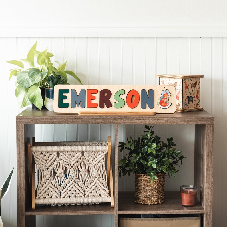 May include: Wooden name puzzle with the name "EMERSON" spelled out in colorful letters. The puzzle is on a shelf with a small plant and a decorative box.