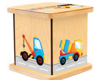 May include: Wooden toy box with a light wood finish. The box has a colorful design of a tow truck and a cement mixer on the front. The box is perfect for storing toys or other small items.