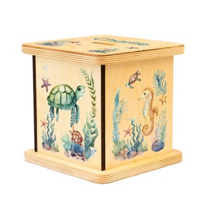 May include: A wooden money box with a slot on top, featuring sea life illustrations. The box is decorated with a green sea turtle, seahorse, starfish, and aquatic plants. The name "Charlie" is written on the top.