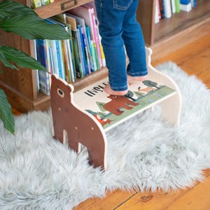 May include: A wooden step stool with a bear-shaped side panel. The top features a forest scene with a bear, fox, and the word "INGLE". The stool is light-colored with brown and green accents, and is placed on a fluffy white rug.
