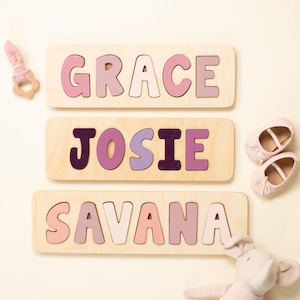 May include: Three rectangular wooden name puzzles featuring the names GRACE, JOSIE, and SAVANA in various shades of pink and purple. A light pink wooden teether and a pair of light pink baby shoes are placed near the puzzles.