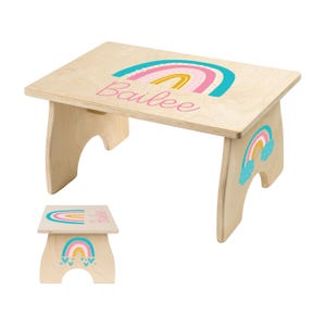 May include: A small, wooden step stool with a rainbow design in teal, pink, and yellow. The name "Bailee" is written in pink script. A smaller version of the stool is also visible. The stool is made of light-colored wood.