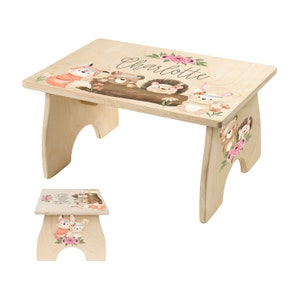 May include: A wooden step stool with a whimsical woodland animal design. The stool is painted with a fox, bear, hedgehog, and bunny wearing flower crowns. The name "Charlotte" is painted on the top of the stool.