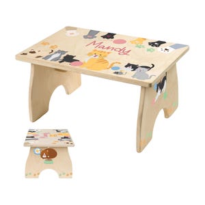 May include: A light-colored wooden step stool featuring a rectangular top and curved legs. The top is decorated with colorful cat illustrations and the name "Mandy" in pink script. Paw prints and other cat-themed designs adorn the stool.