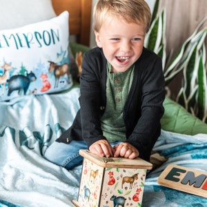 May include: A young child sits on a blue and green blanket, smiling and playing with a wooden box with a woodland animal design. The box has a hinged lid and is decorated with foxes, deer, and bears. The child is wearing a black cardigan and blue jeans. The blanket has a white pillow with the word "EMERSON" printed on it.