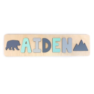 May include: A wooden name puzzle featuring the name AIDEN in alternating mint green and grey block letters. The puzzle includes a dark grey bear silhouette on the left side and a dark grey mountain range silhouette on the right side.