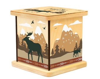 Op de afbeelding: Houten spaarpot met een berg- en elandontwerp. De spaarpot is lichtbruin met een donkerbruine rand. Het ontwerp toont een eland die voor een bergketen met besneeuwde toppen staat. Er zijn ook kleine bomen en vogels in het ontwerp.