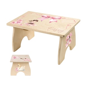 May include: A light-colored wooden step stool featuring a ballerina and floral design. The stool has a rectangular top with a pink bow and the name "Casey" written on it. The sides are decorated with ballet shoes and flowers.