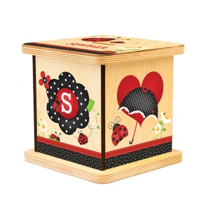 May include: A wooden ladybug-themed box with a light wood finish. The box features ladybug and floral designs, a black and white polka dot pattern, and the name "Sophia" on top. The letter "S" is prominently displayed on the front.