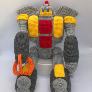 May include: A handmade crochet robot toy in shades of grey, yellow, red, and orange. The robot has a detailed design with wings, a head, and articulated limbs. The toy is made of soft yarn and is suitable for children.