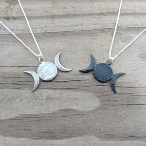 May include: Two necklaces with moon pendant designs. One pendant is silver-toned, the other is black. Each pendant features a central circle with two crescent moons. The necklaces are displayed on a wooden surface.