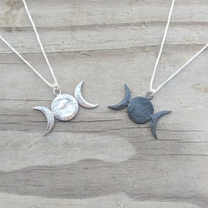 May include: Two silver-toned necklaces with a triple moon pendant design. Each pendant features a central circle with two crescent moons. The necklaces are displayed on a weathered wooden surface, showcasing the celestial-themed jewelry.