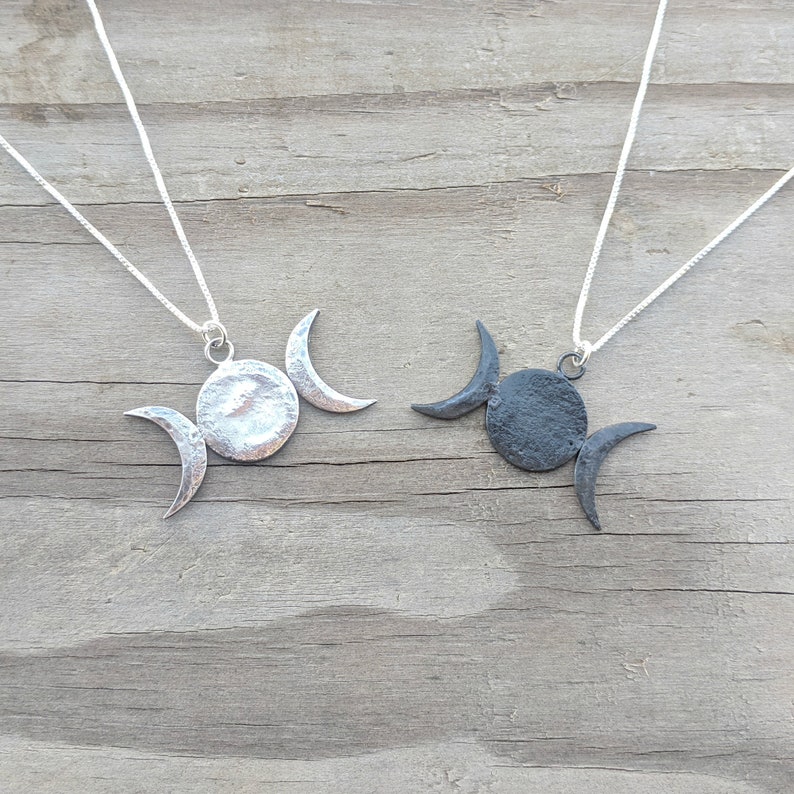 May include: Two silver-toned necklaces with a triple moon pendant design. Each pendant features a central circle with two crescent moons. The necklaces are displayed on a weathered wooden surface, showcasing the celestial-themed jewelry.