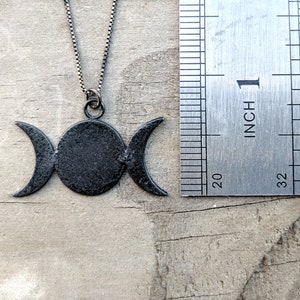 May include: A black metal necklace featuring a triple moon pendant. The pendant has a central circle flanked by two crescent moons. The necklace is shown with a ruler, with the pendant measuring approximately 1 inch in width.