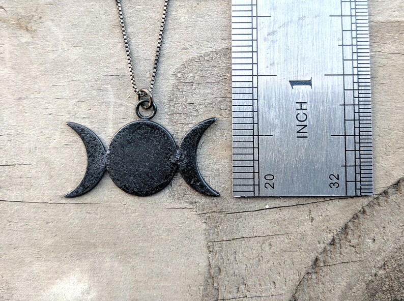 May include: A black metal necklace featuring a triple moon pendant. The pendant has a central circle flanked by two crescent moons. The necklace is shown with a ruler, with the pendant measuring approximately 1 inch in width.