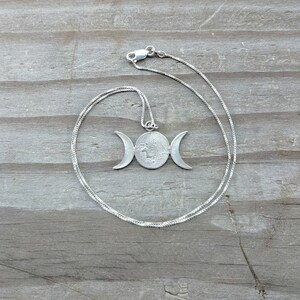 May include: A silver-toned necklace featuring a pendant with a central circle and crescent moons on either side. The pendant hangs from a delicate chain. The necklace is displayed on a weathered wooden surface.