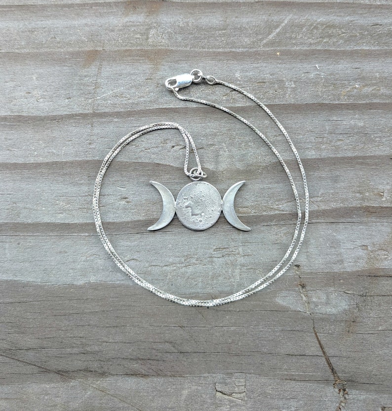 May include: A silver-toned necklace featuring a pendant with a central circle and crescent moons on either side. The pendant hangs from a delicate chain. The necklace is displayed on a weathered wooden surface.
