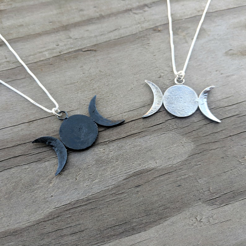 May include: Two silver-colored necklaces with a triple moon pendant. One pendant is black, the other is silver. Each pendant features a central circle with two crescent moons on either side. The necklaces are displayed on a wooden surface.