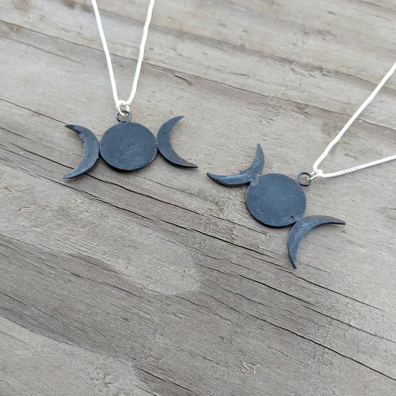 May include: Two dark gray metal necklaces featuring a triple moon pendant design. Each pendant has a central circle with crescent moons on either side. The pendants hang from thin, white cords, set against a weathered wooden surface.