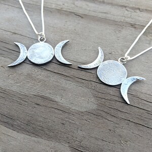 May include: Two silver-toned necklaces featuring a triple moon pendant design. Each pendant has a central circle flanked by crescent moons. The pendants hang from delicate silver chains, set against a weathered wooden surface.