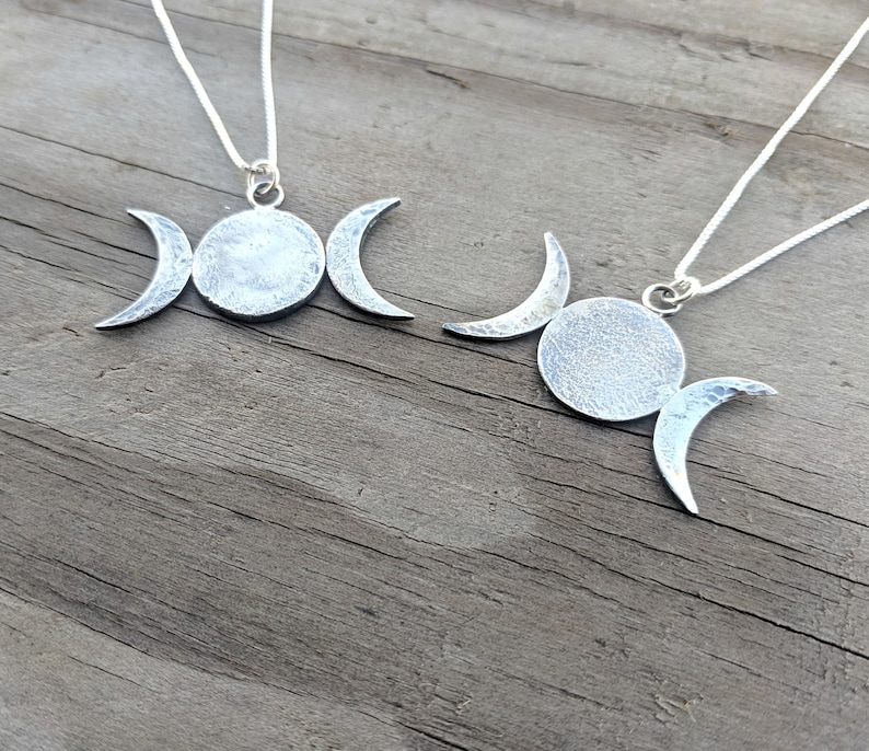 May include: Two silver-toned necklaces featuring a triple moon pendant design. Each pendant has a central circle flanked by crescent moons. The pendants hang from delicate silver chains, set against a weathered wooden surface.