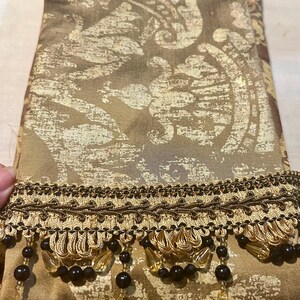 May include: A decorative fabric with a gold and brown damask pattern. The fabric is trimmed with a dark brown and gold fringe, accented with black beads and clear teardrop-shaped crystal pendants.
