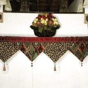 May include: A red, gold, and brown patterned valance with tassels hangs above a white wall. The valance is made of fabric with a leopard print and a paisley design.