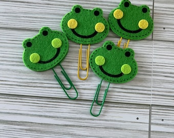 May include: Four green felt frog-shaped paper clips with yellow eyes and black stitched smiles. The clips are on a light wood surface.