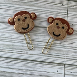 May include: Two brown felt monkey head paper clips with gold metal clips. The monkeys have brown ears, brown fur, and a tan face with black eyes and a stitched smile.