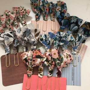 May include: A set of 10 gold paper clips with floral fabric bows. The bows are in various floral patterns, including pink, blue, and green. The paper clips are on a pink, brown, and blue background.