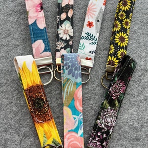 May include: Seven colorful fabric keychains with floral patterns. Each keychain has a silver metal ring attached. The keychains are arranged in a fan shape on a gray surface.