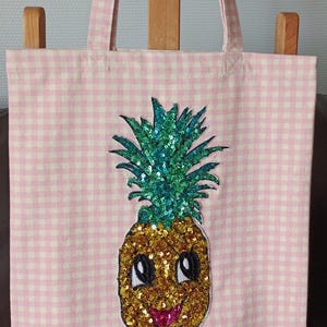 May include: Pink gingham tote bag with a smiling pineapple design. The pineapple is embellished with gold and teal sequins. The bag has matching pink handles and is made of a light fabric.
