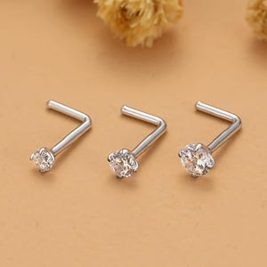 May include: Three silver L-shaped nose studs with clear, round gemstones. The jewelry is displayed on a light brown surface with dried flowers in the background. The studs are small and delicate, with a simple, elegant design.