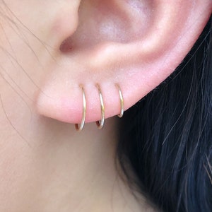 May include: Three small, gold-coloured hoop earrings are worn in a person's ear. The earrings are evenly spaced and appear to be made of a thin metal. The skin around the ear is visible, with a soft, natural tone. The background is blurred.